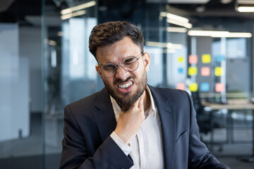 Fototapeta premium A businessman in glasses grimaces while touching his sore throat. He is in an office setting, possibly experiencing an illness.