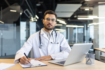 A handsome doctor in a white coat sits at his desk, writing on a clipboard with a laptop in front...