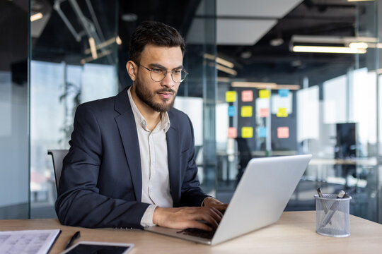 A focused businessman in a suit, working on a laptop at his desk in a modern office environment.