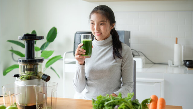 Young Asian woman holding a glass of fresh green juice in a clean, modern kitchen, promoting healthy living and natural wellness.