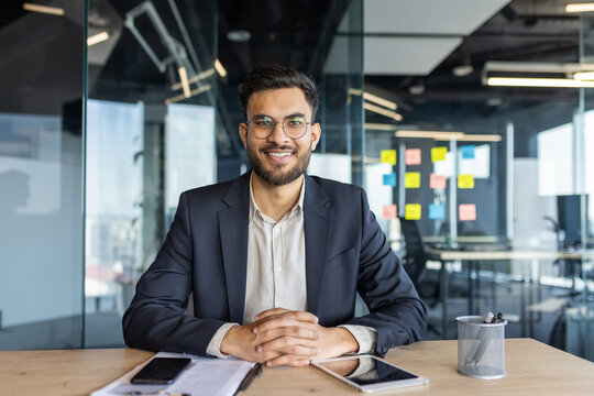 A smiling businessman sits at a desk in a modern office. He wears glasses and a suit, appearing confident and professional.