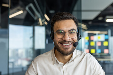 A smiling man wearing a headset, looking directly at the camera in a modern office setting.