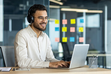 A smiling man wearing headphones uses a laptop in a modern office setting. He has glasses and is...