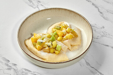 Two pancakes with kiwi and orange slices sprinkled with powdered sugar in bowl