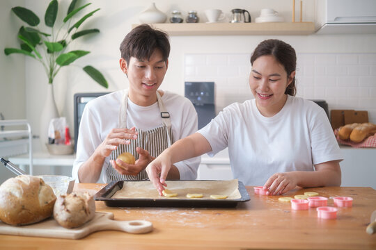Smiling couple placing cookie dough shapes onto a baking tray in a modern kitchen. - Powered by Adobe