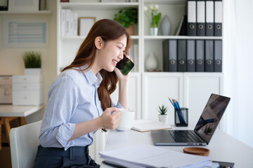 Smiling young woman talking on the phone while holding a coffee cup and working on a laptop in a modern office.