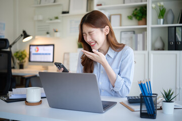 Laughing young woman reading a funny message on her smartphone while working on a laptop in a...