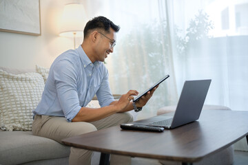 Fototapeta premium Man sitting on sofa smiling while using a digital tablet and laptop for remote work in a bright living room.