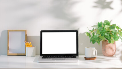 Fototapeta premium Laptop with blank white screen on a white desk, accompanied by coffee mug, and green plant in natural light.