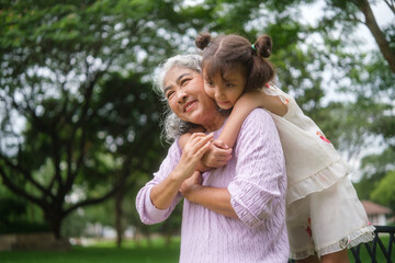 Grandmother smiling warmly while her granddaughter hugs her from behind in a green park.