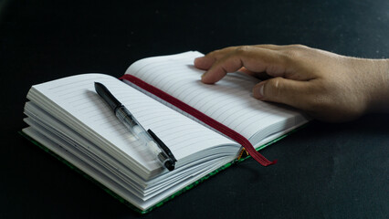 Side view of notebook and pen on book with male hand above, placed on black table against black...