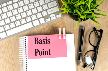Basis point text on pink sticky note clipped to notebook with keyboard, pen, glasses, and plant on...