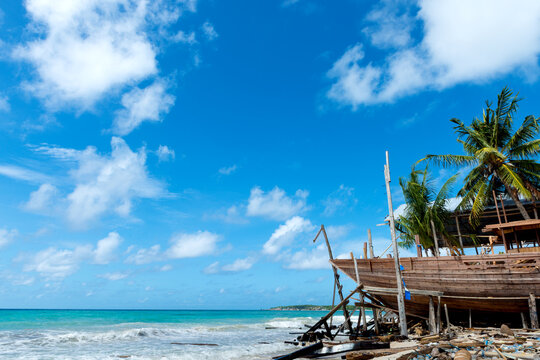 Traditional Wooden Boat Under Construction on Tropical Beach with Palm Trees, Clear Blue Sky, and Turquoise Sea in Remote Coastal Setting