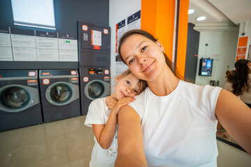 Mother and daughter taking a selfie while waiting in a self-service laundromat, looking at camera...