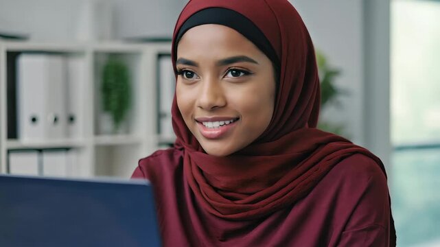 Smiling Muslim woman in maroon hijab talking on video call at office &ndash; natural expressions, modern professional setting, ideal for business, diversity, tech or online communication themes
