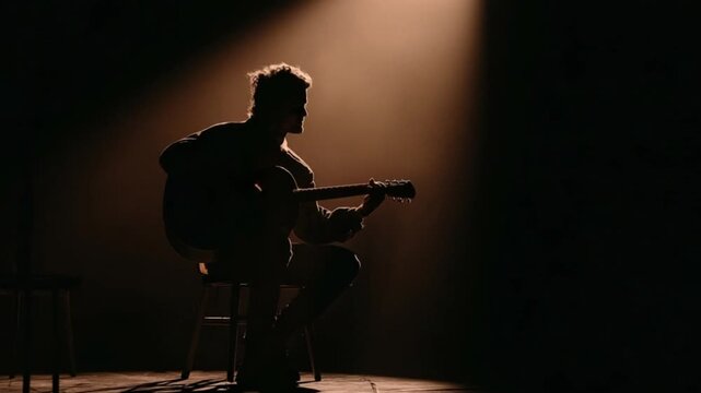 Silhouetted guitarist sits on a stool under a spotlight, playing acoustic guitar
