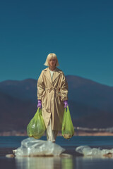 Fototapeta premium A woman is walking on the water, wearing an oversized beige trench coat and purple gloves, holding green bags in her hand with plastic trash inside. 
