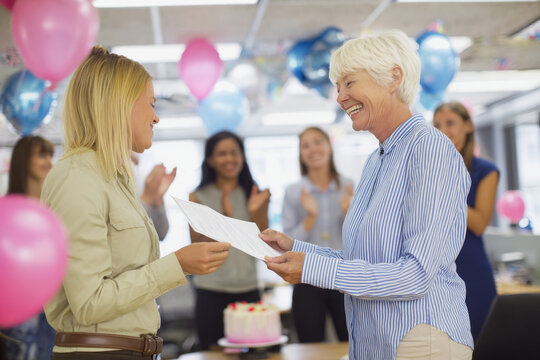 Elderly Caucasian businesswoman smiling as she awards a certificate to a young employee during a staff appreciation event in the break room, surrounded by balloons, cake, and applause.
