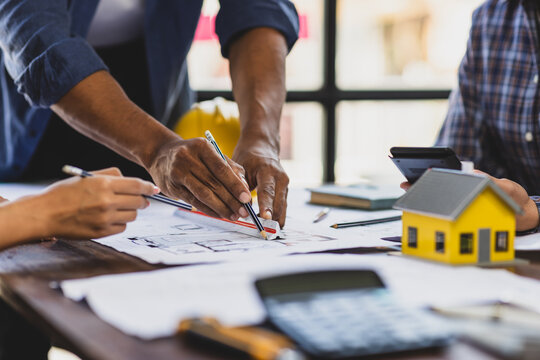 Architects and engineers are pointing at building plans and discussing over a model house, collaborating on a construction project.