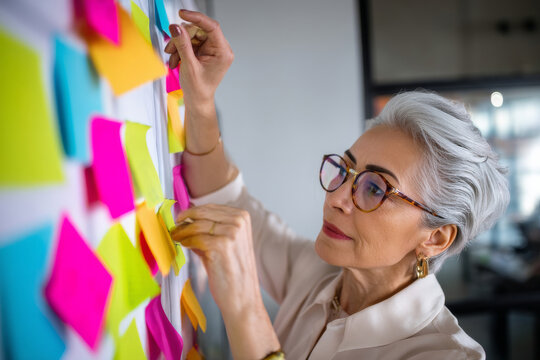 Hispanic elderly businesswoman with silver hair organizing colorful sticky notes on a project wall, planning a product launch timeline with sharp focus and an experienced eye for structure.