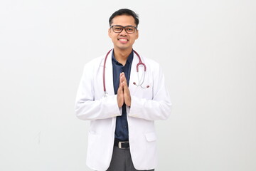 Fototapeta premium Asian adult male doctor standing smiling, wearing doctor's coat, stethoscope around neck, holding hands on chest while saying namaste, greeting gesture, looking at camera