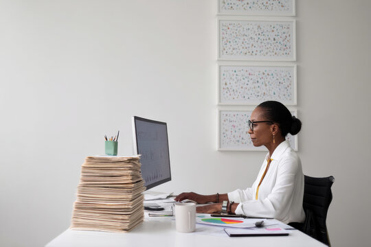Mature Black business woman reviewing quarterly numbers on a desktop monitor, sitting in a minimalist executive office, with a stack of printed reports beside her and motivational artwork on the white - Powered by Adobe