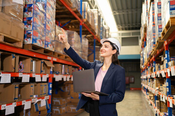 A confident female supervisor in a suit and hard hat inspects stocked shelves in a large warehouse.