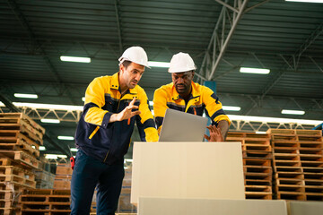 Two Warehouse Workers in hard hats review inventory data on a laptop amid stacked pallets and shelving in a modern distribution center.