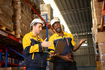 Two Warehouse Workers in hard hats review inventory data on a laptop amid stacked pallets and shelving in a modern distribution center.
