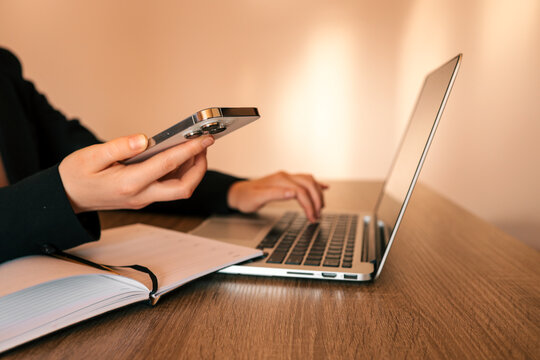 Person holding smartphone while typing on laptop keyboard on wooden desk with notebook nearby, warm natural light, working or browsing online - Powered by Adobe