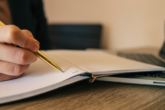 Close-up of hand writing notes in open planner next to laptop keyboard on wooden desk in office