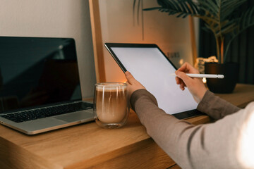 Person using digital tablet with stylus near laptop and glass of latte on wooden desk in cozy workspace