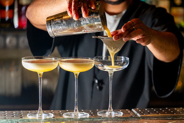 Bartender pouring a yellow cocktail through a strainer into a coupe glass, with two other cocktails...