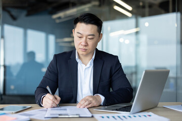 Serious and focused young Asian man sitting in the office at a table with a laptop and working with documents