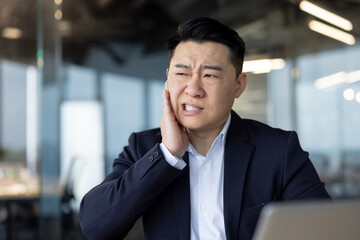 Close-up photo of an Asian businessman working in an office, holding his cheek with his hand, feeling a severe toothache