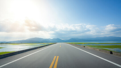 Fototapeta premium Empty asphalt road leading towards distant mountains under a bright blue sky with scattered clouds