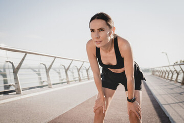 Fototapeta premium Strong Caucasian woman female runner in earphones activewear taking a moment break to rest after her morning run outdoors in city bridge landscape. Wellness lifestyle concept, copy space