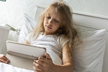 A young girl looks sad and focused while interacting with a tablet device while lying in her bed in...