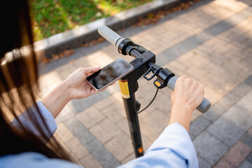 Woman gripping handlebars of electric scooter while interacting with smartphone. Urban pavement...