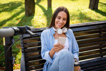 Close up of woman sitting on a park bench with headphones holding smart phone using internet mobile...