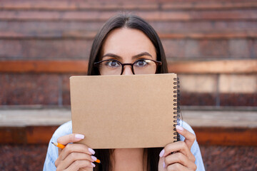 Funny young female student in eyeglasses holding a notepad with both hands, covering her face. Space for advertisement. Concept of college study programs, academic courses, and learning classes.