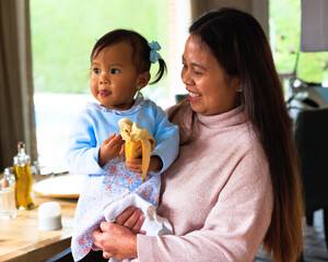 Happy moment with loving mother holding toddler eating banana in dining room