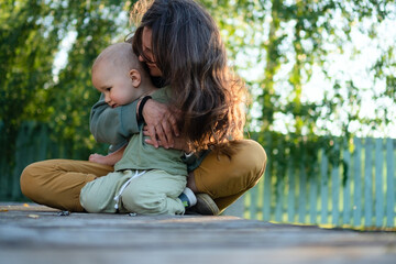 Mother and child sitting on a wooden surface in a garden setting, surrounded by greenery. The...