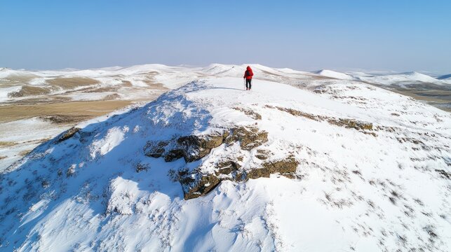A lone hiker in a red jacket stands atop a snow-covered mountain ridge, overlooking a vast, pale-yellow landscape