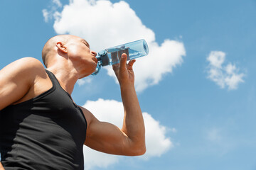 Athlete sun-tanned man wearing black sleeveless vest top drinking water against sky. Concept water...