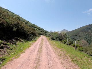 rural road winds through verdant hills, Cordel - Experience rural Spain in Cueto Cordel, Cantabria. A winding path meets a road under a cloudy sky surrounded by lush greenery.