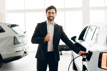 Salesman showing thumbs up while charging electric car in dealership