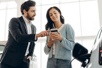 Car salesman showing smartphone app to woman buying electric vehicle