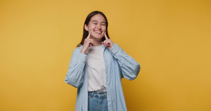 smiling young woman playfully points to her own cheeks, expressing joy and whimsy on vibrant yellow background