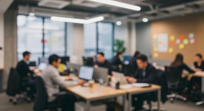 A blurred view of a modern open-plan office, with a diverse team of professionals working on laptops at shared desks.